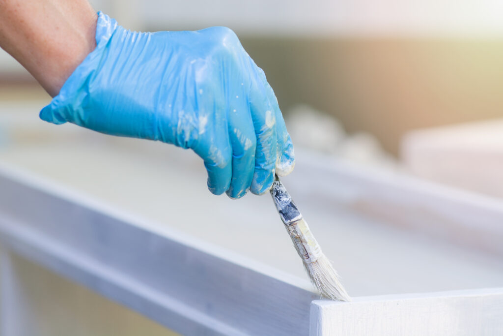 Hands in blue gloves hold a paintbrush and paint a new wooden plank with white paint outdoors. The man makes repairs. Close-up. The concept of construction, renovation, self-repair. Everything to Know Before Painting Cabinets
