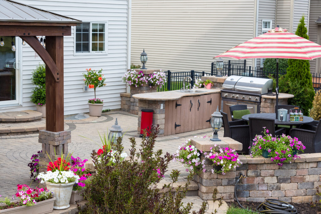 Summer flowers around an upscale brick patio with wooden gazebo, outdoor kitchen with BBQ and comfortable wicker dining furniture. outdoor countertop material